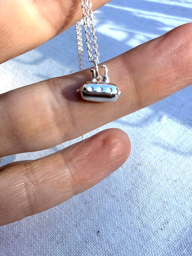 Silver necklace with a submarine pendant held between fingers on a light background

