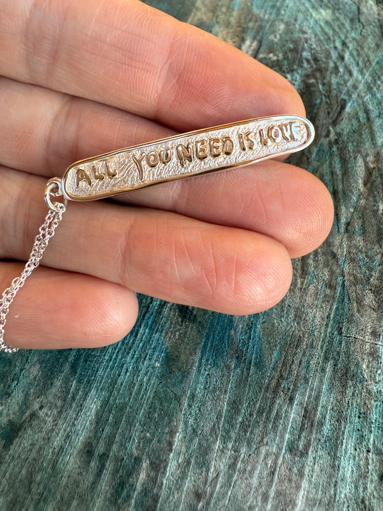 Silver necklace with 'All You Need Is Love' pendant held by a hand on a textured surface