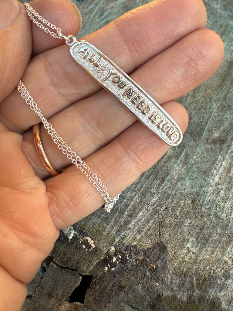 Silver necklace with a bar pendant with All You Need is Love carved on it held by a hand on a wooden surface