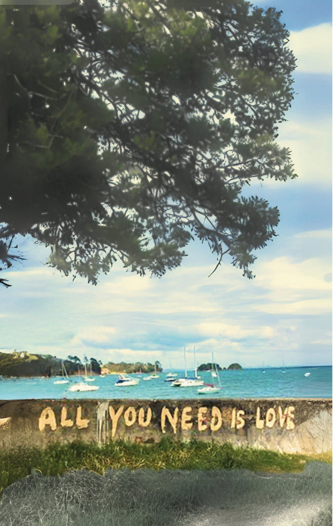 Mural with 'All You Need Is Love' on a wall by a waterfront with trees and boats on Waiheke Island, NZ.