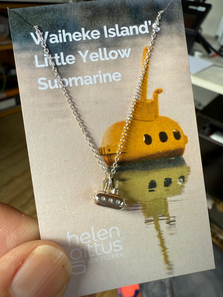 Silver submarine necklace on a card with 'Waiheke Island Little Yellow Submarine' text, held by a hand.

