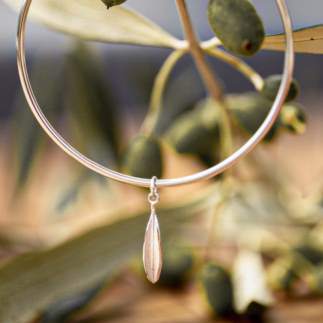 Small sterling silver olive lead charm on plain sterling silver bangle pictured among olive leaves and olives.