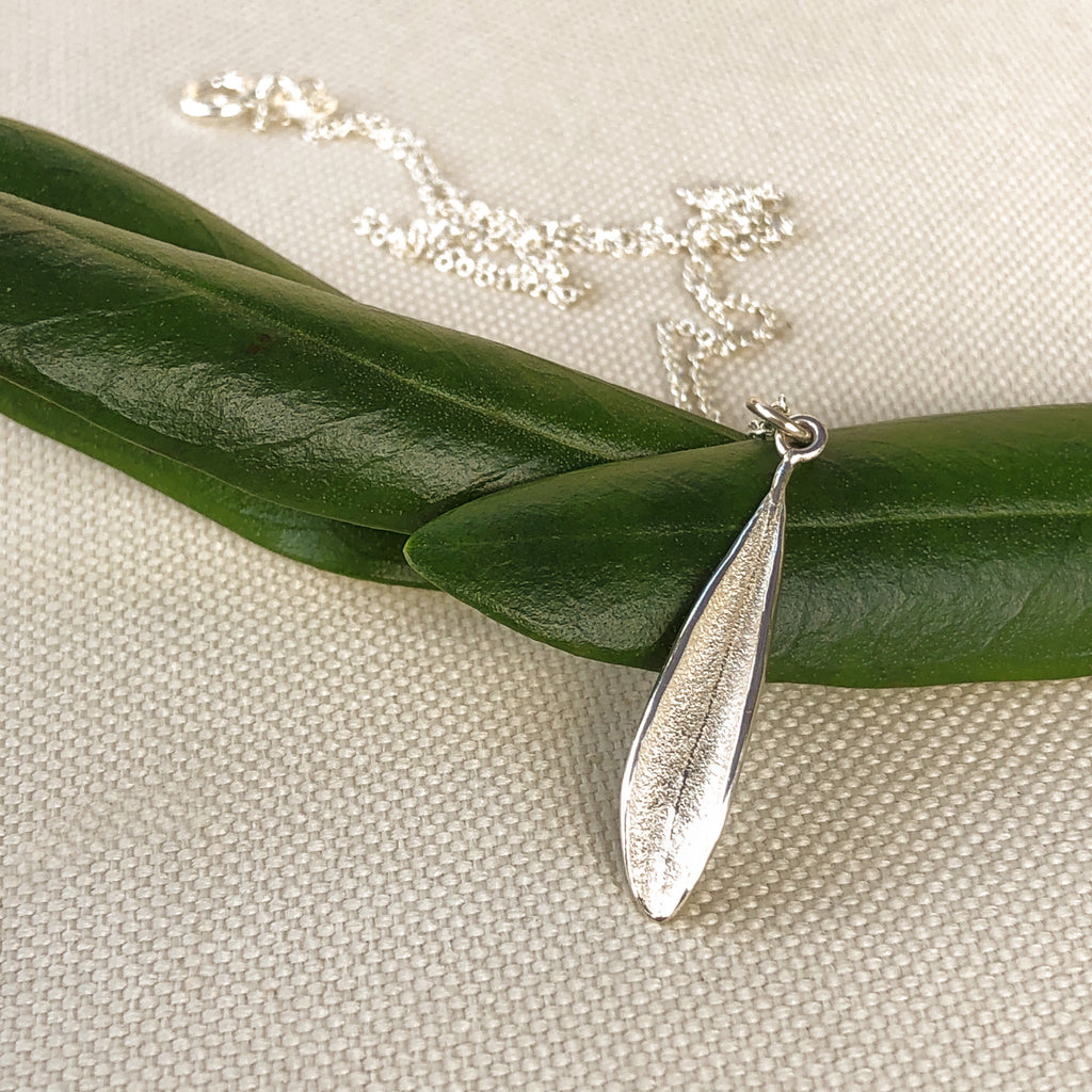 Close up of a handmade sterling silver picual olive leaf pendant on fine sterling silver cable chain resting on olive leaves.